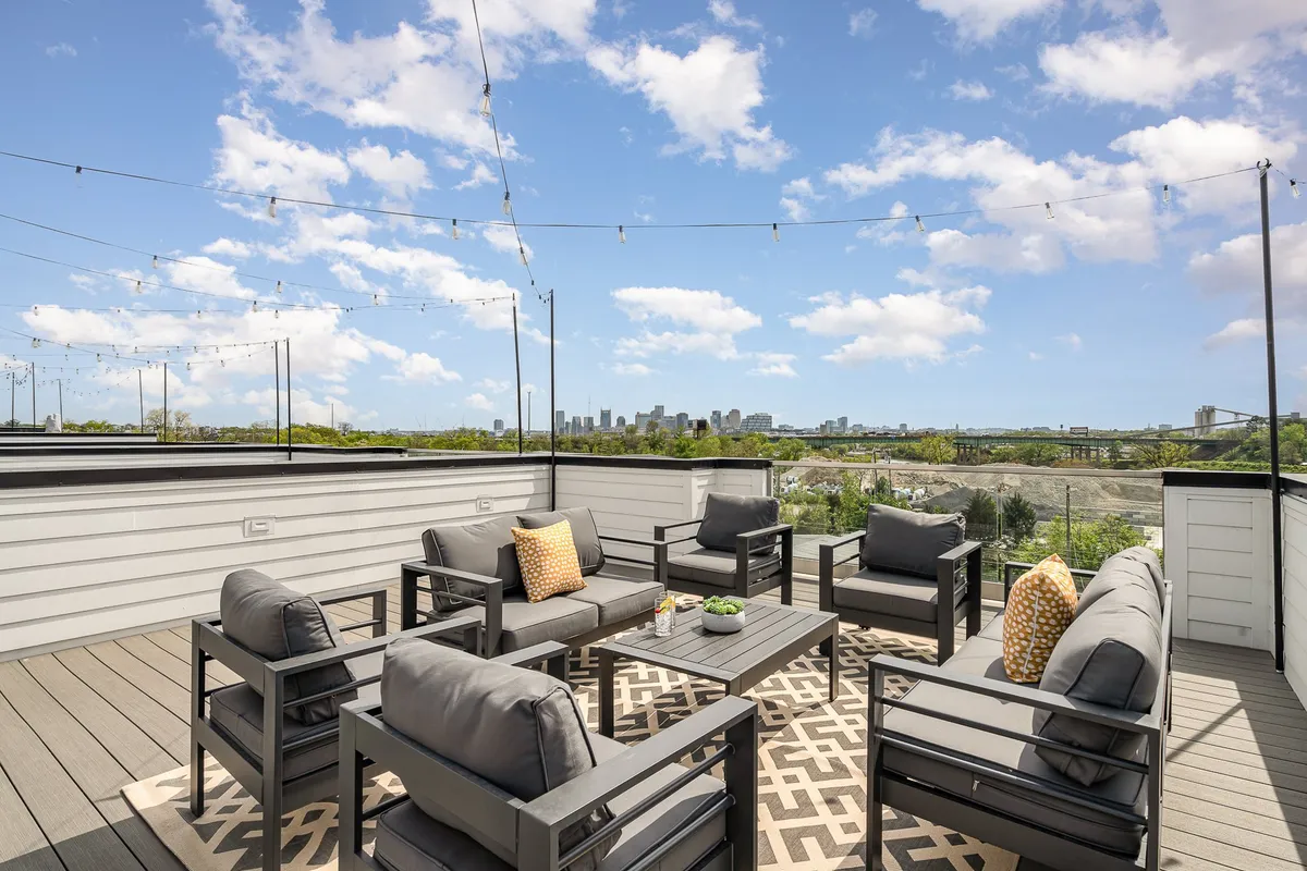 The photo presents a stylish outdoor seating area on a rooftop, accented with lush plants on the coffee table, decorative string lights, and sweeping views of the city skyline. The ambiance is casual and cheerful, suggesting opportunities for socializing or enjoying the view.