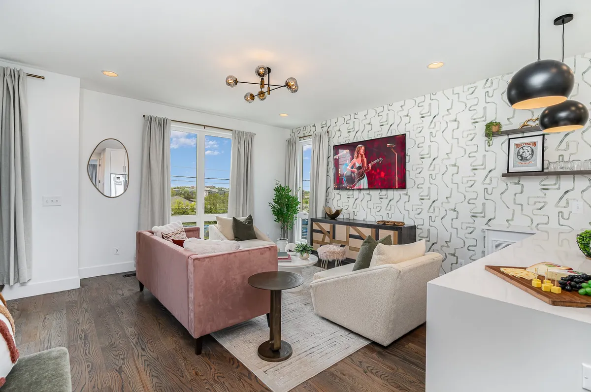 A stylish living room featuring a vibrant pink sofa and a white armchair, set against geometric wallpaper. Large windows fill the space with natural light, and a TV displaying artwork creates a cozy focal point. The setup is perfect for relaxing, entertaining guests, or watching a movie together.