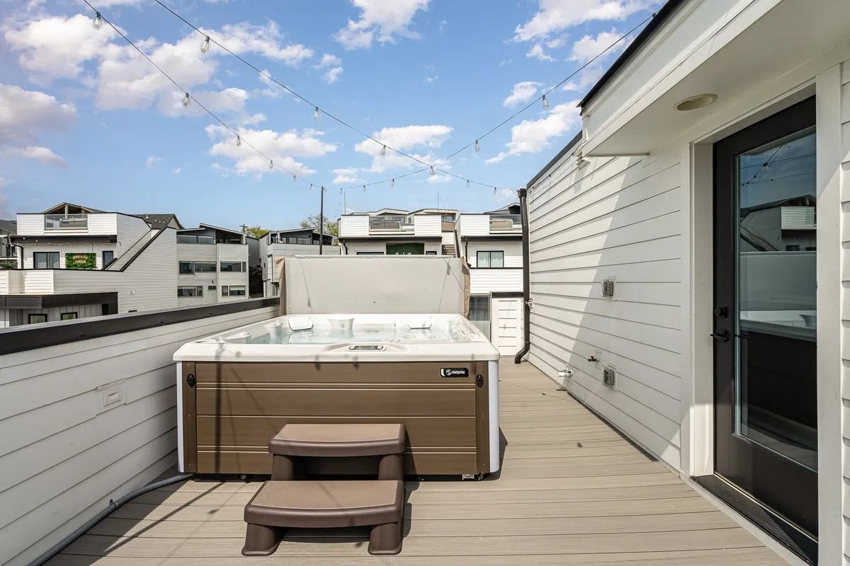 An inviting rooftop deck with a hot tub, cozy string lights, and comfortable seating provides an intimate setting for groups to relax and socialize outdoors. The evening ambiance is perfect for unwinding after nights out, with the city skyline in the distance and an atmosphere ideal for sharing stories under the stars.