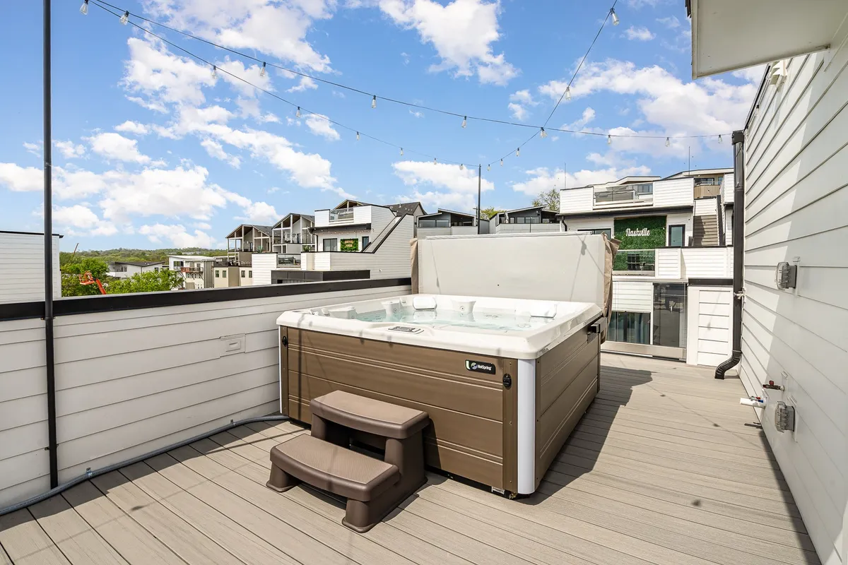 This image captures an inviting rooftop deck at dusk featuring a bubbling hot tub, cozy deck area, and warm string lights overhead. The relaxing setup is ideal for both social gatherings and tranquil evenings, with the city skyline visible in the distance.