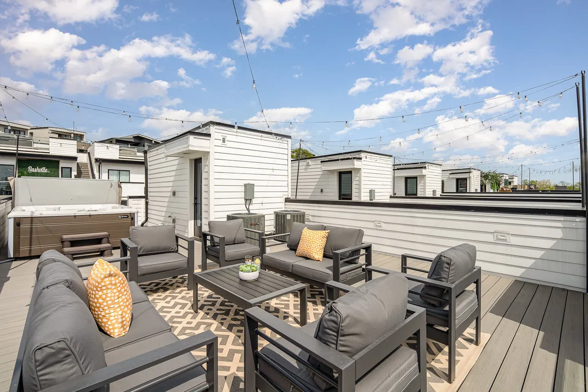 A welcoming rooftop space is depicted with a hot tub, plush outdoor seating, colorful decorative pillows, and string lights overhead. The setup is perfect for both celebratory occasions and quiet moments, with the cityscape beyond providing an atmospheric backdrop.