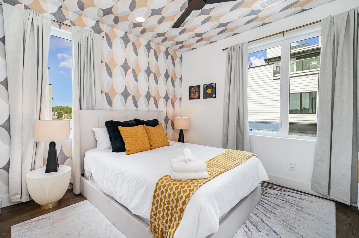 A bright and stylish bedroom featuring a bed with colorful cushions, geometric patterned wallpaper, and a large window that allows natural light to flood the space. The atmosphere is serene and visually stimulating, ideal for relaxation and personal retreat.