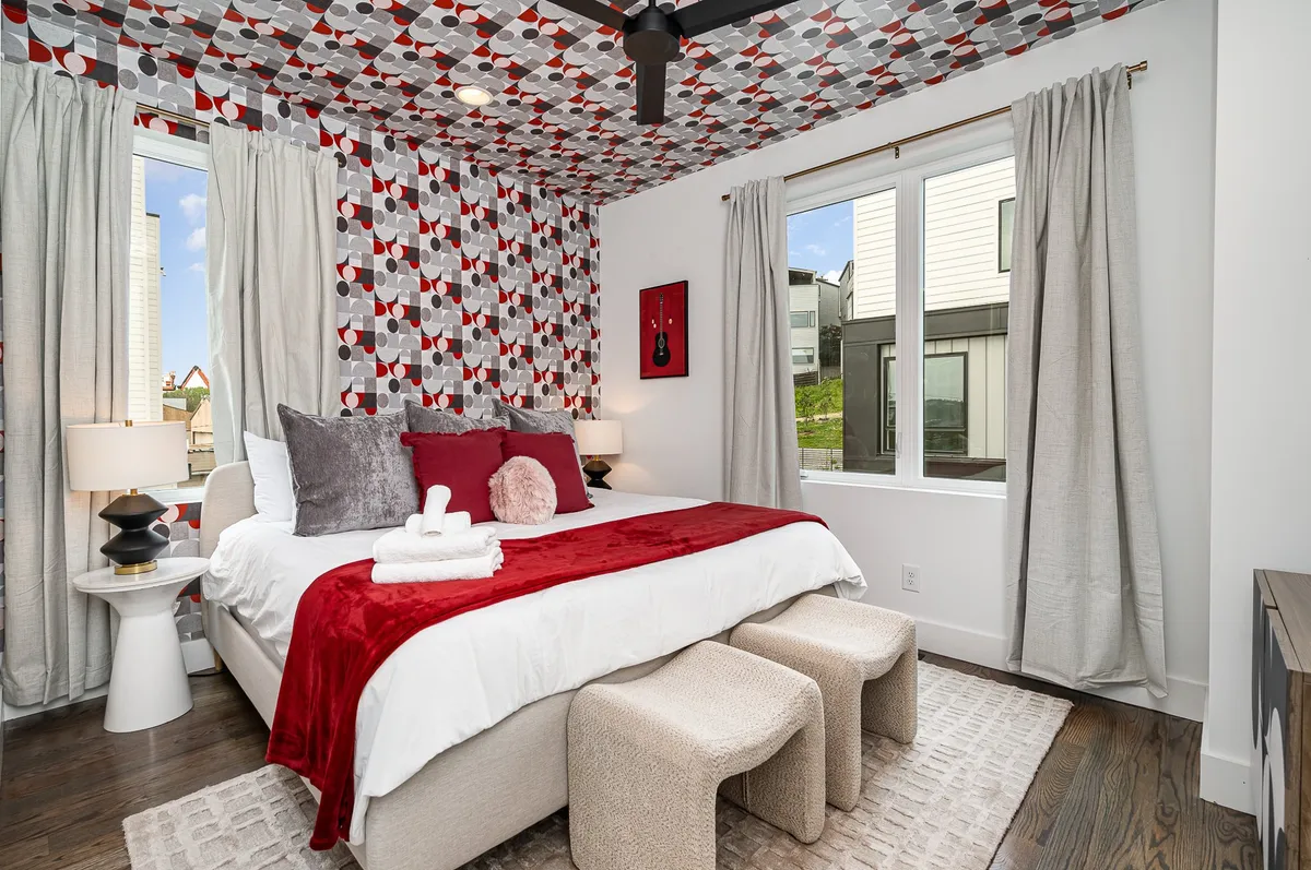 A bedroom with bold red and white bedding, colorful patterned wallpaper, decorative pillows, bedside lamps, and a unique art piece. The space is well-lit and inviting, perfect for unwinding or socializing.