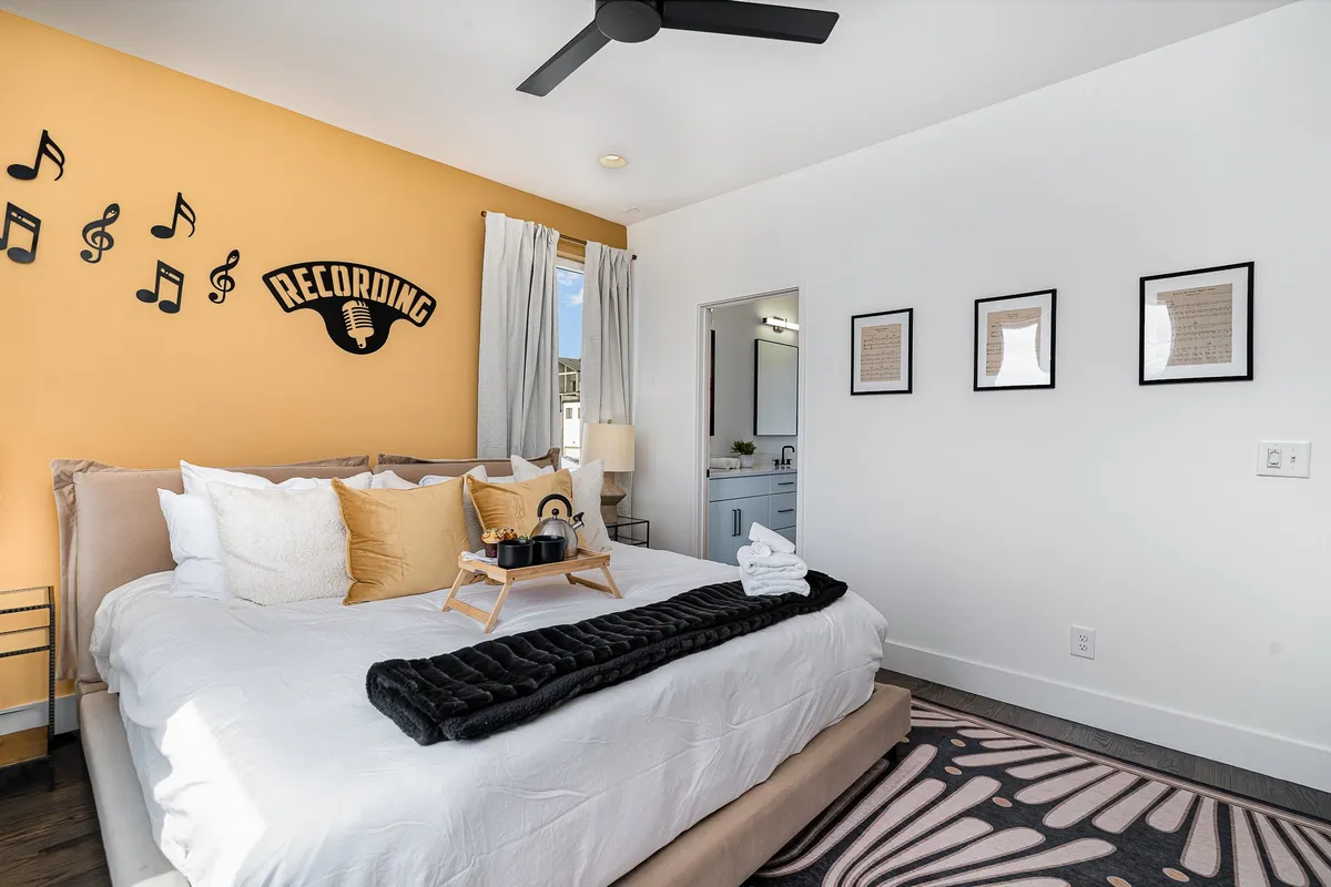 A bedroom with a comfortable bed adorned with decorative pillows, music-themed wall art, and a window with curtains. There's an entrance to a bathroom visible, suggesting en-suite or Jack-n-Jill access, and the overall mood is creative yet restful.
